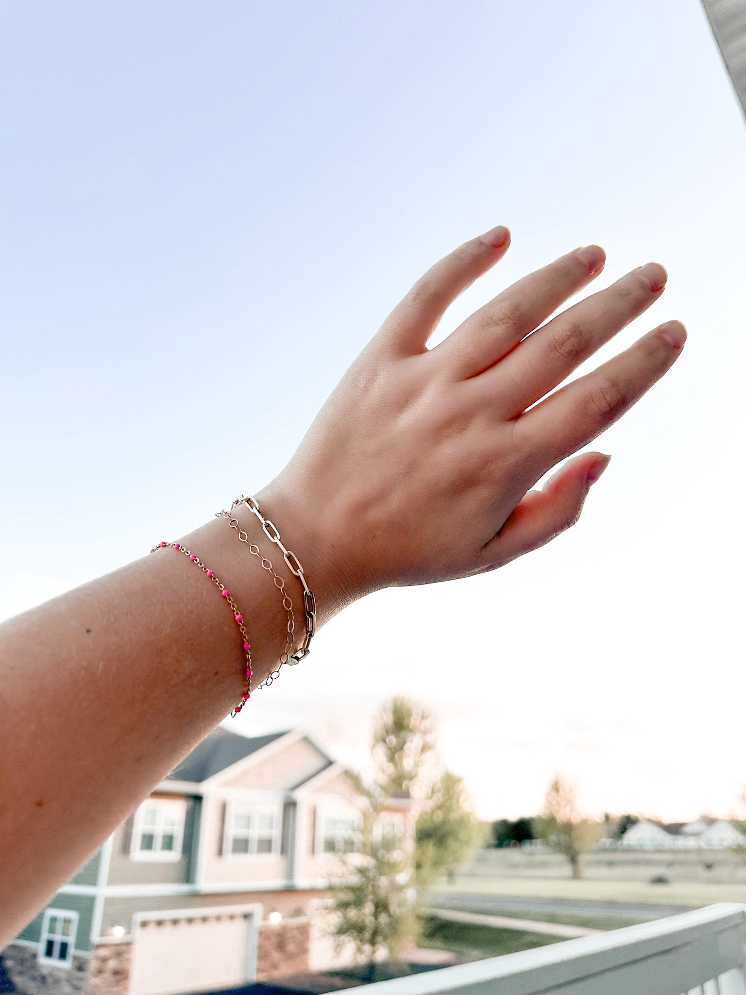 Three stacked permanent bracelets on a wrist — gold-filled rolo, paperclip chain, and a pink gemstone accent